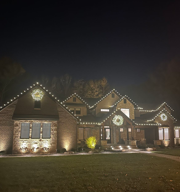 Large stone home with warm white roofline lights and wreaths at night